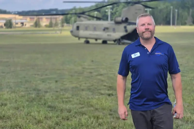 Suburban Bolt President Eric Peterson stands in front of a military Chinook helicopter on a grassy field.
